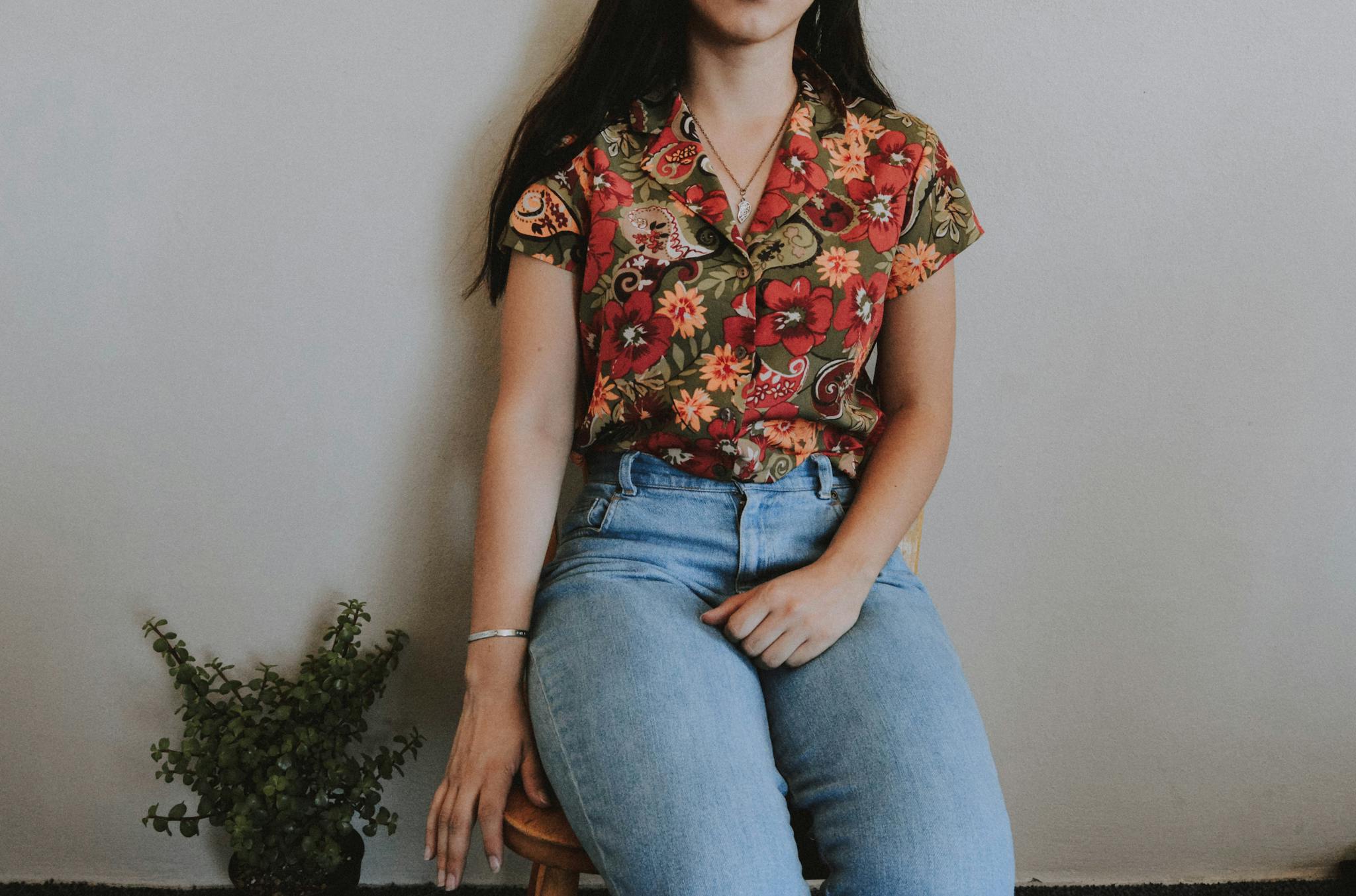 Woman sitting on stool wearing floral shirt and jeans indoors with a plant by her side.